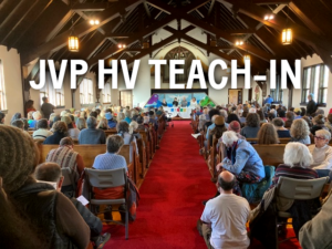 Photo of people sitting in a church, all seats full and extra seating in back, 4 people at a panel in front. Text "JVP HV Teach-in"